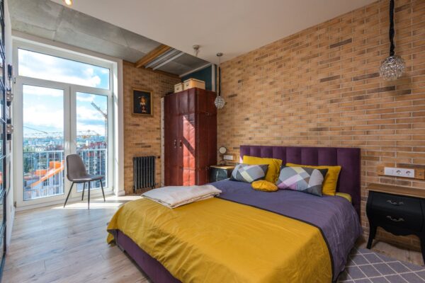 Bright contemporary bedroom with purple and yellow decor, brick wall, and cityscape view.